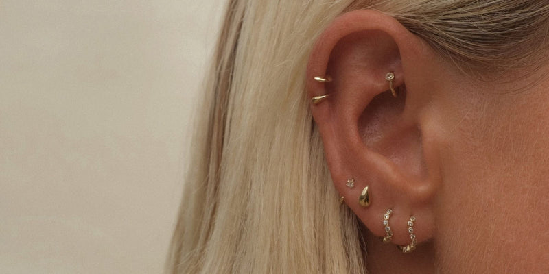 Close-up of an ear with two gold curved flat back hoops stacked on the helix, a diamond hoop in the conch and diamond studs and hoops through the lobe.