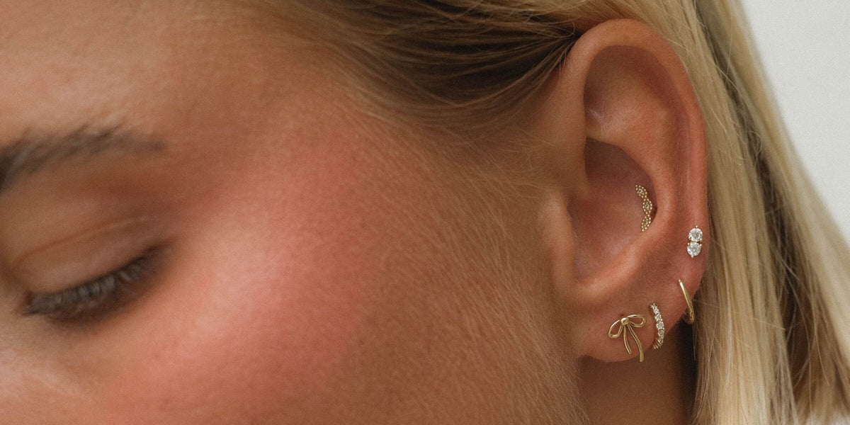 Close-up of an ear with an Escha contour stud in the conch, a double diamond stud, a bow stud and gold hoops through the lobe.