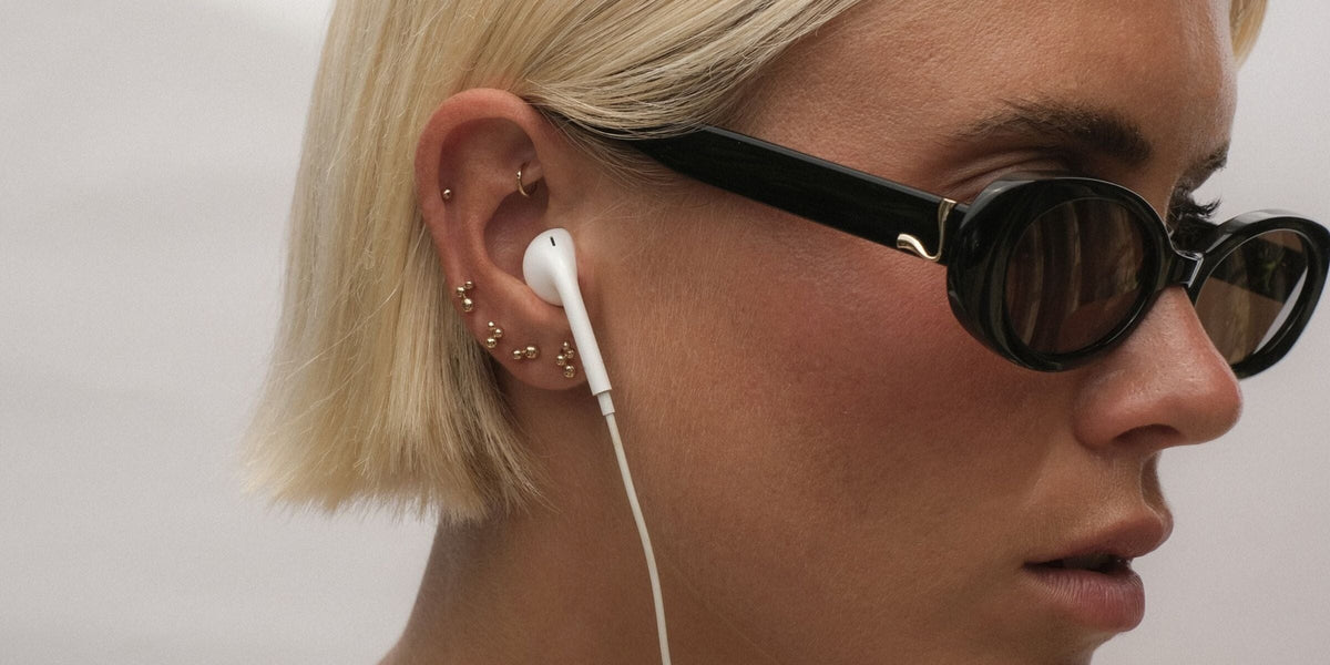 Close-up of an ear stacked with gold flat back studs, worn with wired earphones and sunglasses.