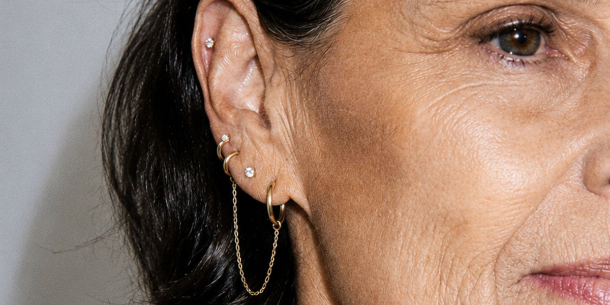 An ear with a stack of gold earrings: diamond studs, huggies and a hoop with a fine chain.