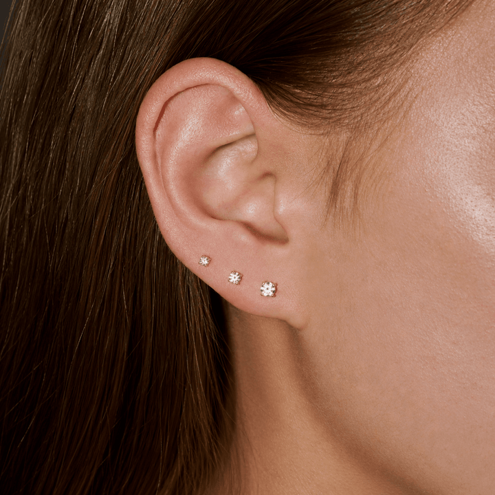 A close-up of a person's ear wearing three small stud earrings in ascending sizes. The earrings appear to be silver-toned with clear stones, set against the person's brown hair and skin.