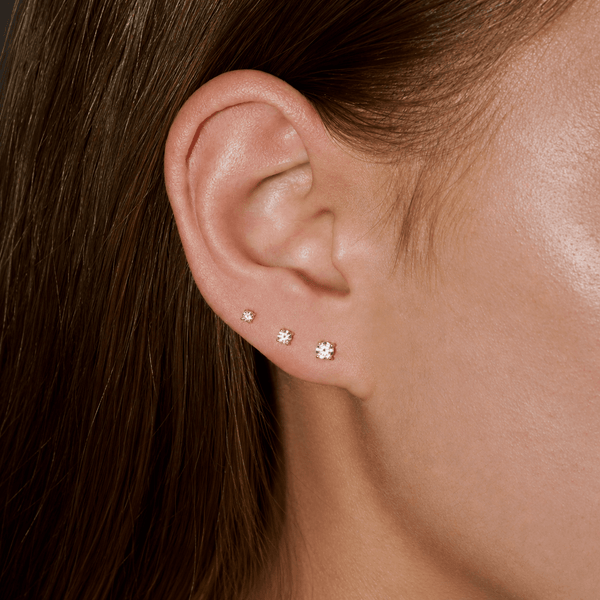 A close-up of a person's ear wearing three small stud earrings in ascending sizes. The earrings appear to be silver-toned with clear stones, set against the person's brown hair and skin.