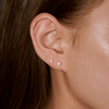 A close-up of a person's ear wearing three small stud earrings in ascending sizes. The earrings appear to be silver-toned with clear stones, set against the person's brown hair and skin.